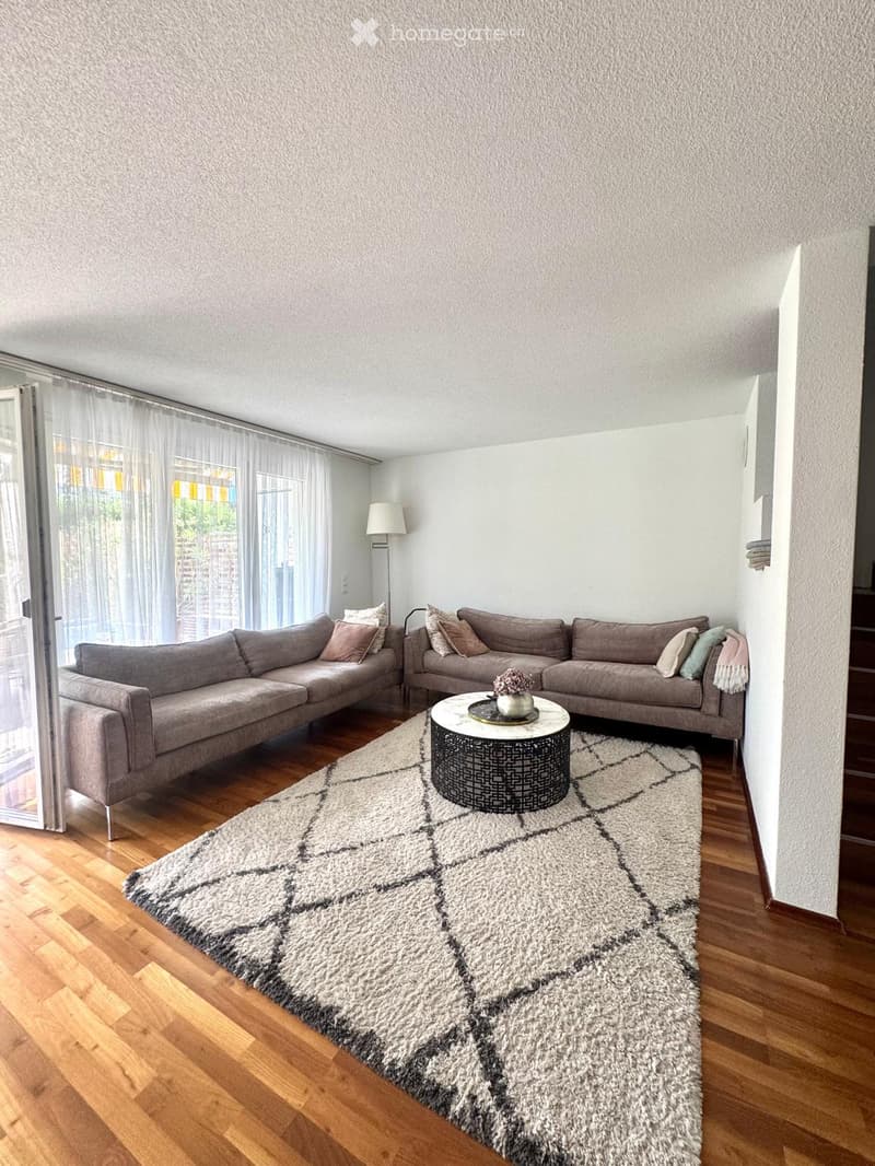 Modern living room with light-colored walls, wooden flooring, and large windows with sheer curtains. Features a grey sectional sofa, a round coffee table with a decorative centerpiece, a patterned rug, and a floor lamp.