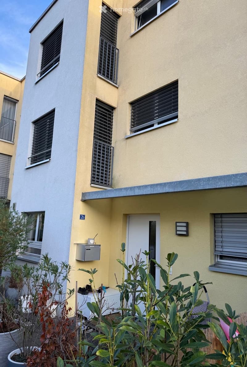Two-story residential building with yellow and grey exterior, multiple windows with blinds, and a small garden area with potted plants in front of the entrance.