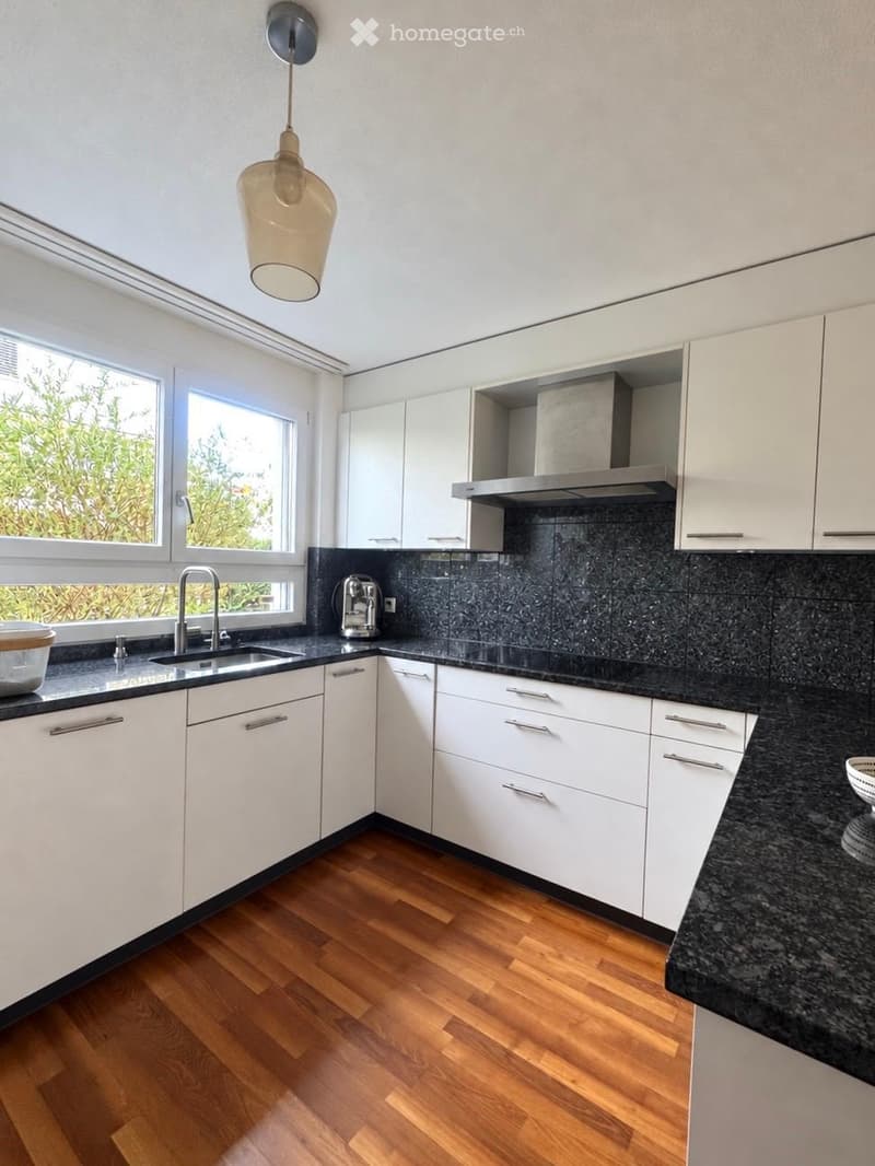 Modern kitchen with white cabinets, black granite countertops, wooden flooring, and a hanging light fixture.