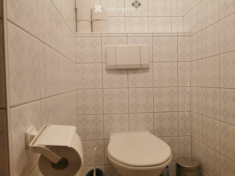 A modern bathroom with a toilet, toilet paper holder, toilet paper, toilet brush, and a small trash can. The walls are tiled with a diamond pattern in light beige.