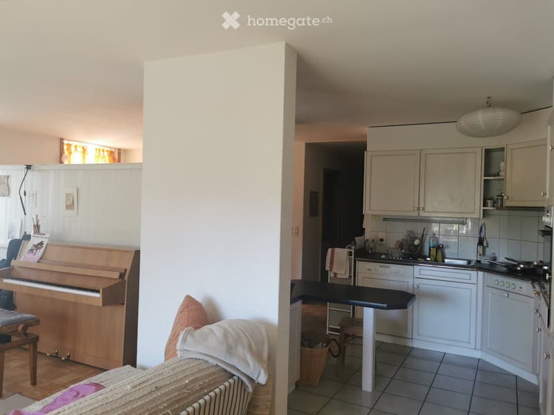A furnished room with a piano, sofa, and a connected kitchen area with cabinets, stove, sink, and oven.