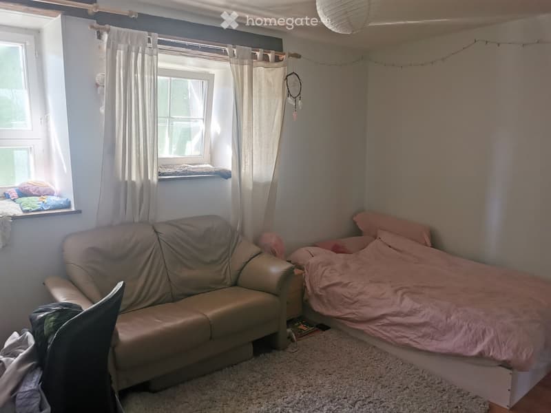 Cozy bedroom with a bed, sofa, chair, and a window with curtains. The room features a soft, light-colored carpet and a small shelf on the windowsill with folded blankets.
