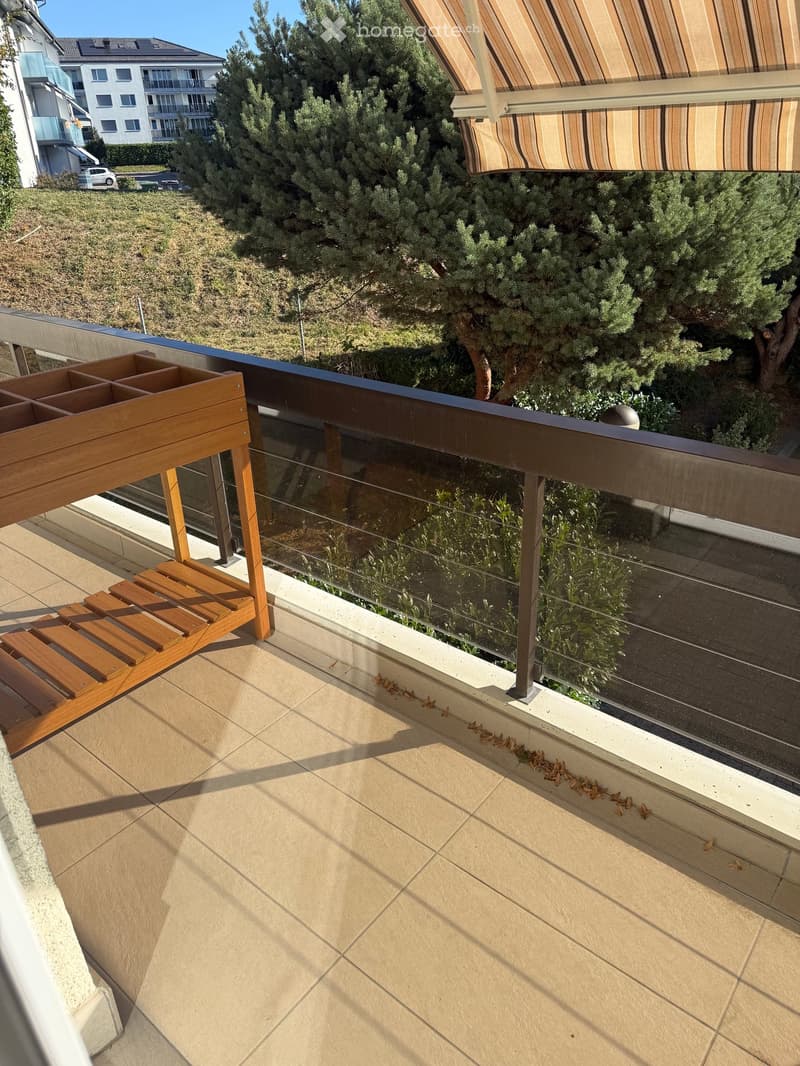 Balcony with tiled floor, glass railing, wooden bench, and outdoor furniture. View of greenery and neighboring buildings.