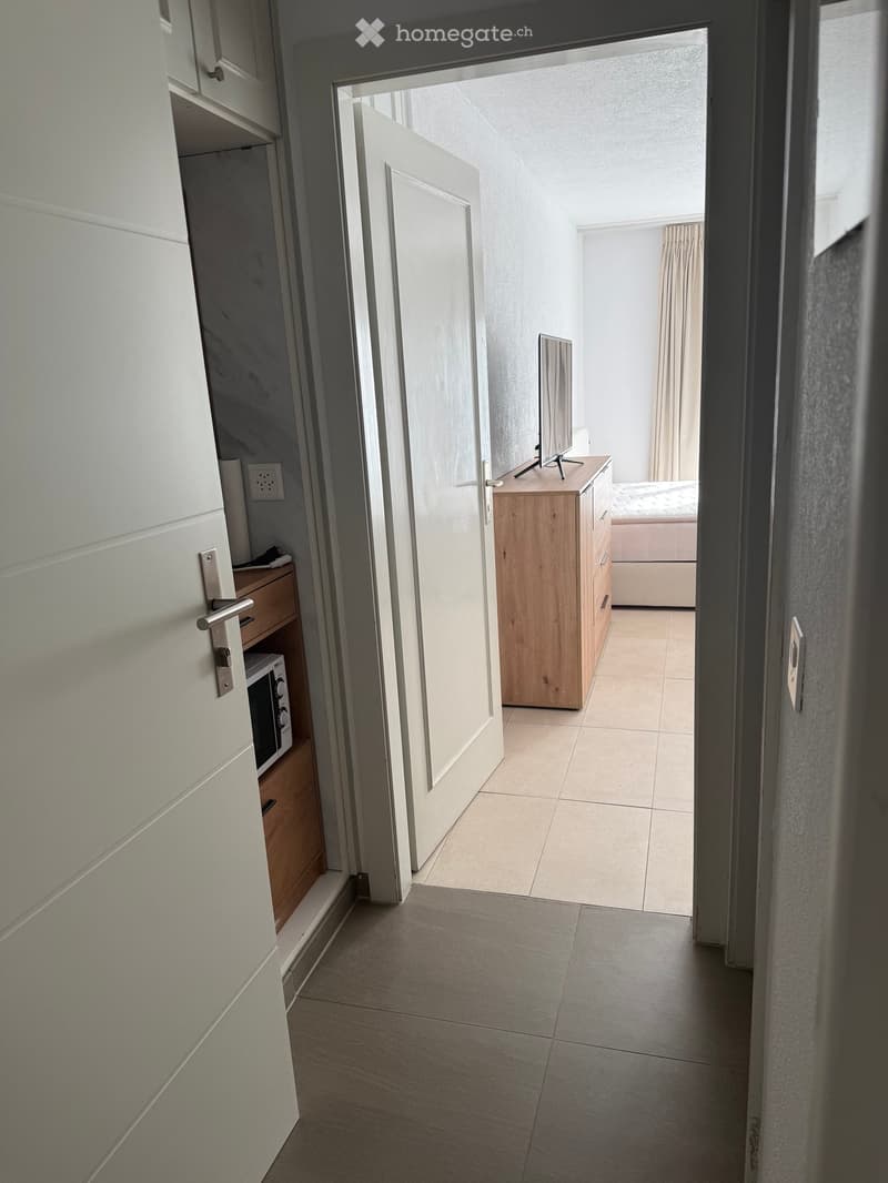 A narrow hallway with tiled flooring, white walls, and a partially open door leading to a bedroom with a bed, dresser, and TV stand.