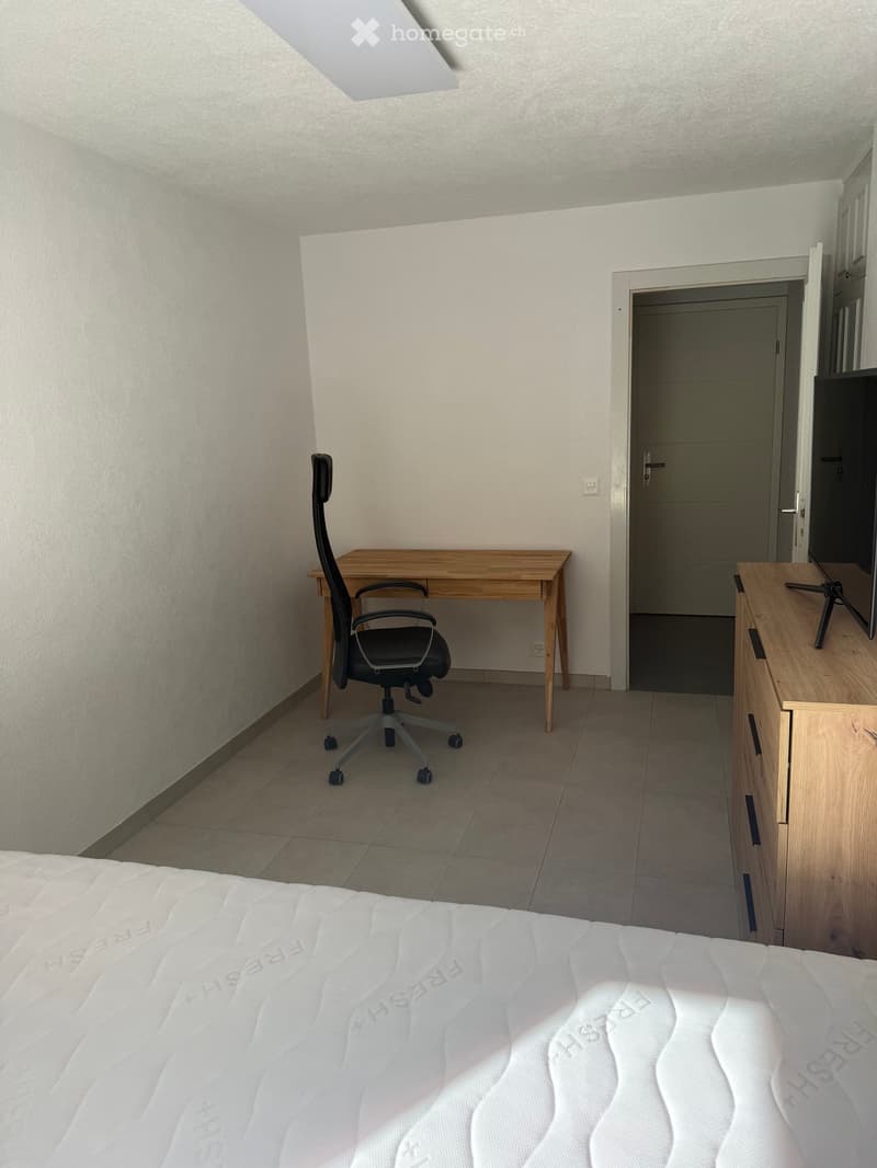 Modern bedroom with a white bed, wooden desk, office chair, TV stand, and TV. The room features light-colored walls and tiled flooring.