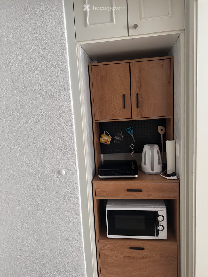 Compact kitchen area with wooden cabinets, microwave, electric kettle, and various small appliances and utensils.