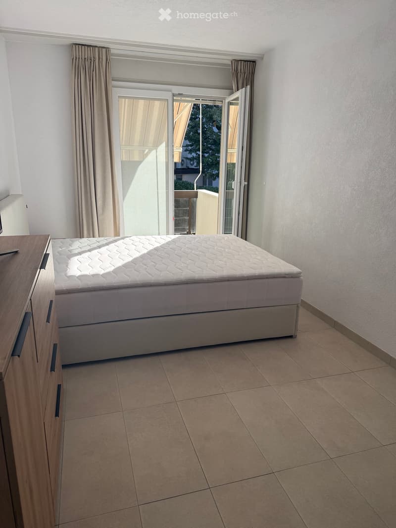 Modern bedroom with a large bed, nightstand, and large window with curtains leading to a balcony.