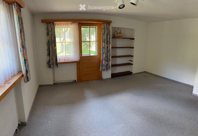 Empty room with carpeted floor, wooden door, window with curtains, built-in shelving unit, and a small decorative item on the shelf.