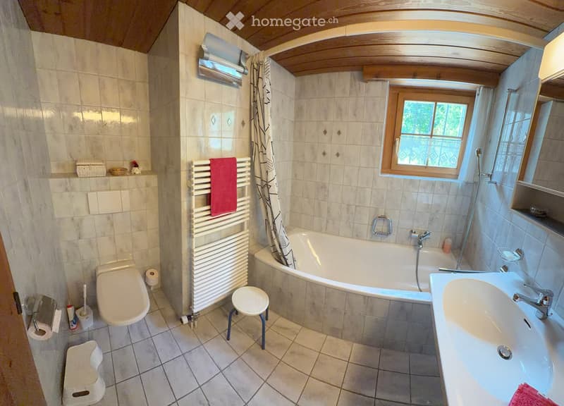 Bathroom with a toilet, bathtub, sink, towel rack, and a small stool. The room features tiled walls and a window with a curtain.