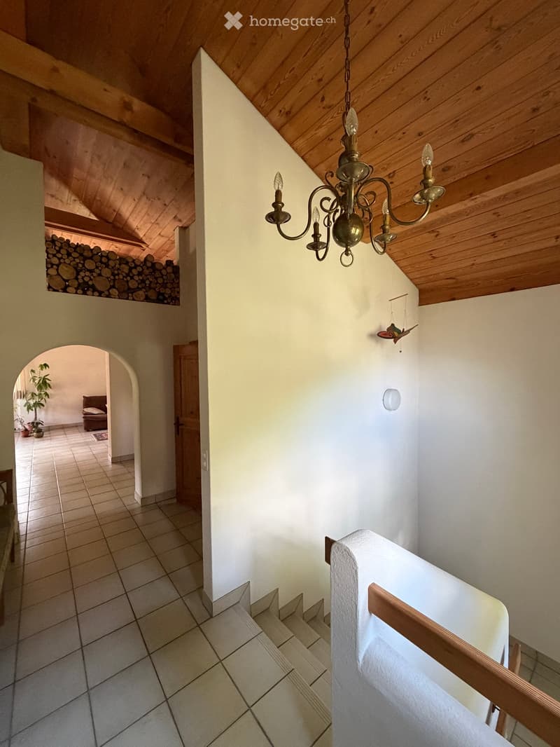 Staircase with wooden railing, chandelier, and visible storage of firewood in the background.