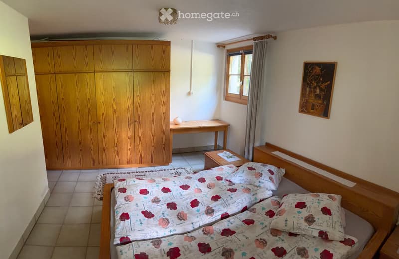 Cozy bedroom with a double bed, wooden wardrobe, small desk, and a nightstand. The room features tiled flooring and a window with curtains.