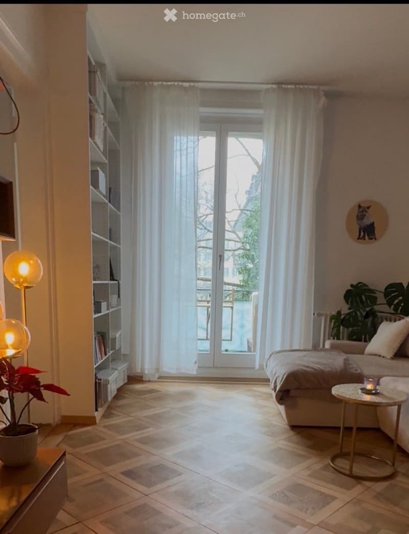 Bright living room with large windows and balcony access, featuring a sofa, coffee table, bookshelf, floor lamp, and decorative plants.