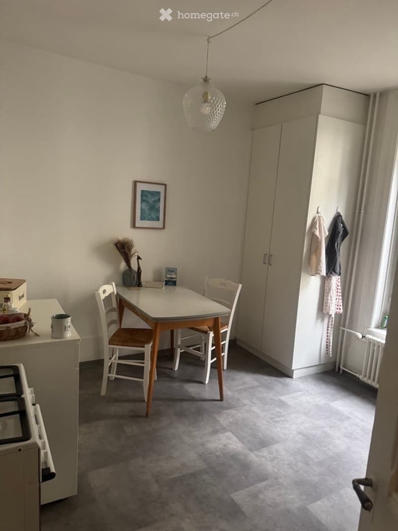 A small, furnished room with a dining table, chairs, a white cupboard, a picture frame on the wall, and a hanging light fixture.