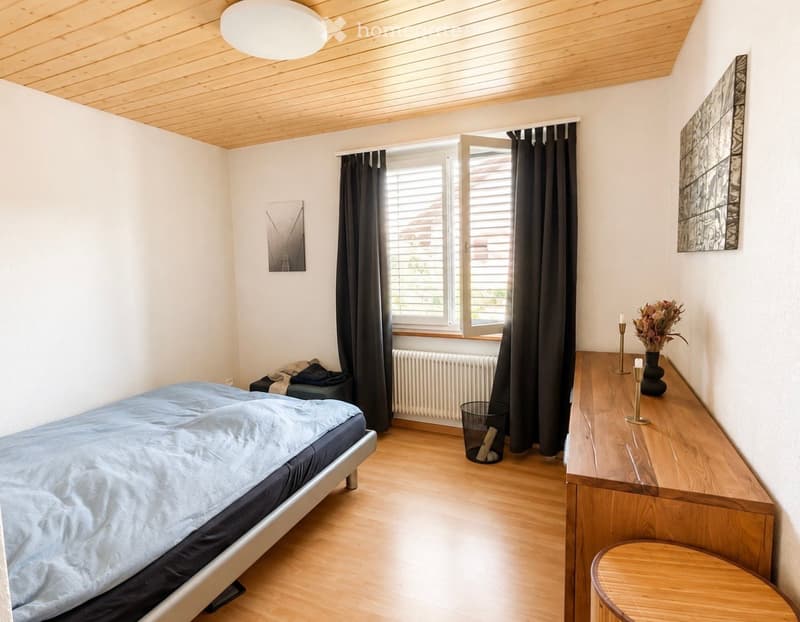 Cozy bedroom with a wooden bed, nightstand, and a small stool. The room features a large window with dark curtains, a radiator, and a trash can near the window. The walls are painted white, and the ceiling has a wooden finish.