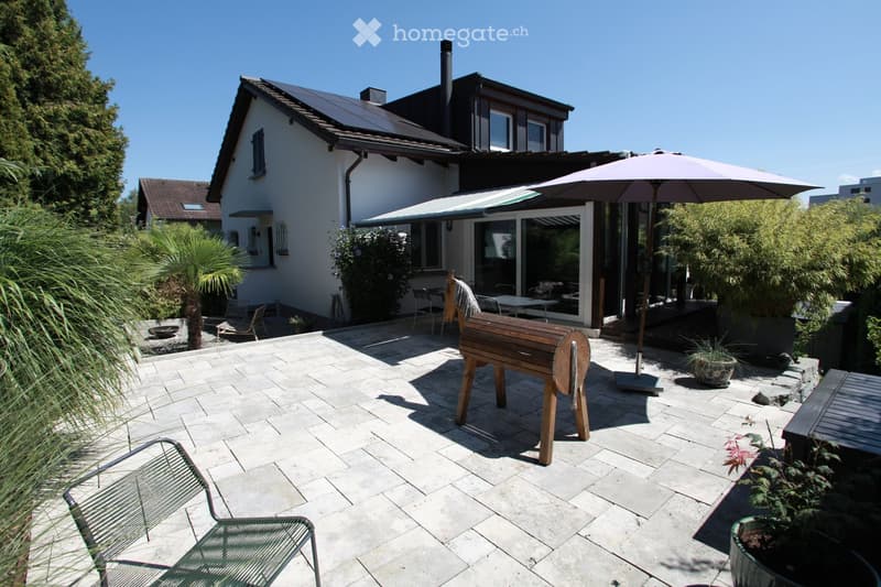 A well-maintained garden patio area with a stone-tiled floor, outdoor furniture including chairs and a table, a large umbrella, potted plants, and a modern house with large windows and a covered patio area.