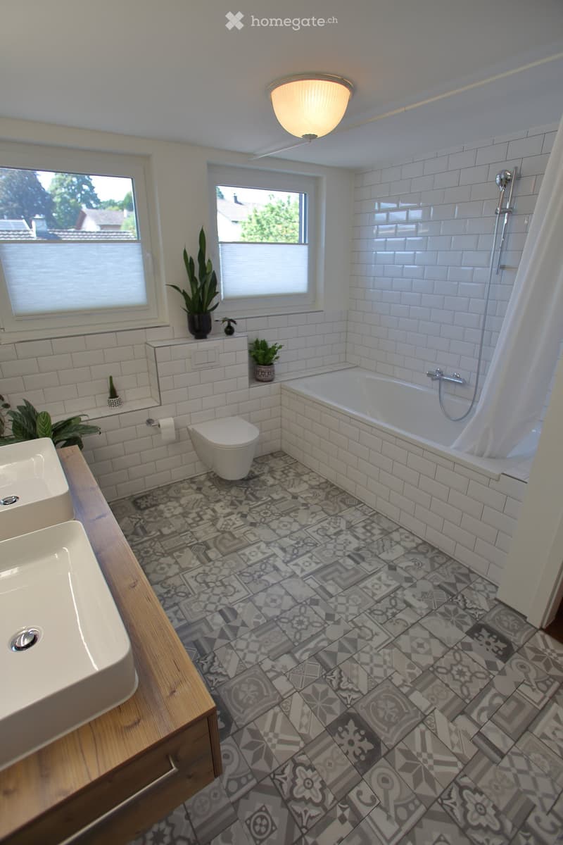 Modern bathroom with white tiles, a double sink, toilet, bathtub with shower, and decorative patterned floor tiles.