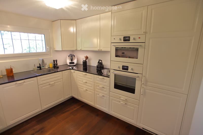 A modern kitchen with white cabinets, a black countertop, and stainless steel appliances including a double oven, sink, and various small appliances.
