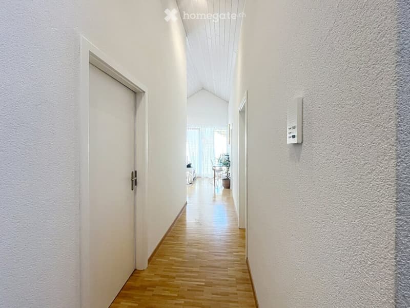 A clean, well-lit hallway with wooden flooring, white walls, and a door on the left leading to another room.