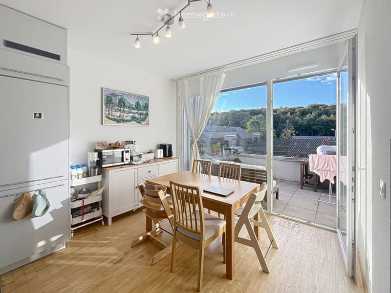Bright dining area with wooden table and chairs, modern appliances, large windows with a scenic view, and a balcony accessible through sliding doors.