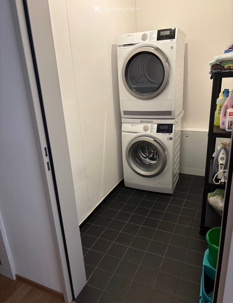 A compact bathroom with a stacked washing machine and dryer unit, a shelf holding cleaning supplies, and an ironing board in the corner.