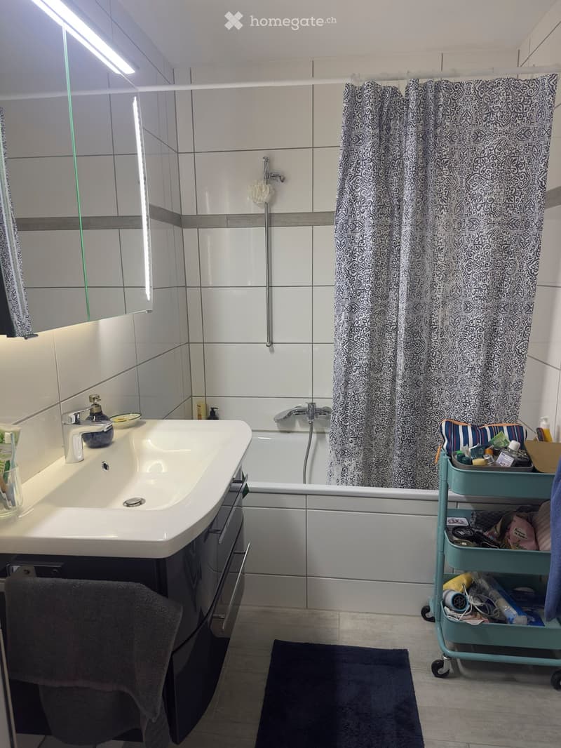 Modern bathroom with white tiles, a sink with a faucet, a bathtub with a shower curtain, a mirror, a towel, and a cart with toiletries.