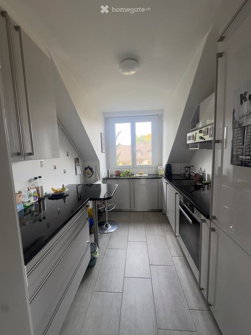 Modern kitchen with stainless steel appliances, grey cabinets, and a black countertop. Features include a refrigerator, oven, sink, and a small bar area with a stool.