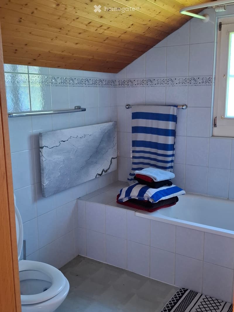 Bathroom with white tiled walls, a toilet, a bathtub, and a towel rack with towels. The room features a marble-like decorative panel and a window providing natural light.