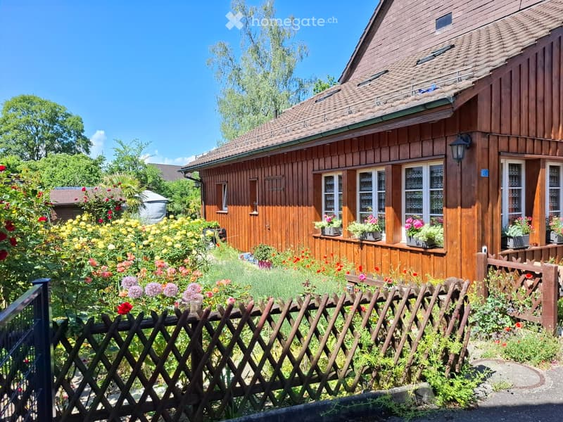 A wooden house with a sloped roof, surrounded by a garden with colorful flowers and a wooden fence.