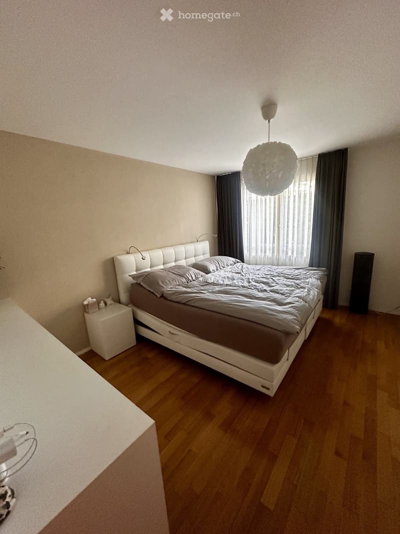 Modern bedroom with a large bed, nightstand, and hanging lamp. Wooden flooring and large window with curtains.