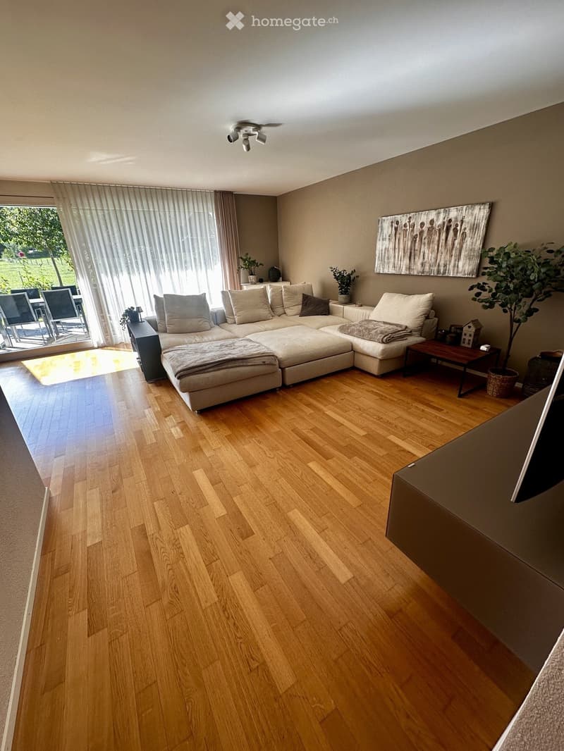 Modern living room with light wooden flooring, large sectional sofa, coffee table, and large windows with sheer curtains.