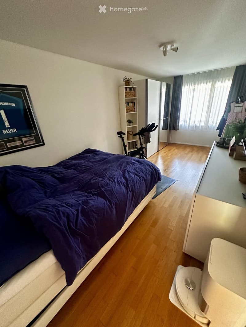 A modern bedroom with a wooden floor, a large bed with dark blue bedding, a wardrobe, a shelf with books and plants, an exercise bike, and a small table with a mirror.