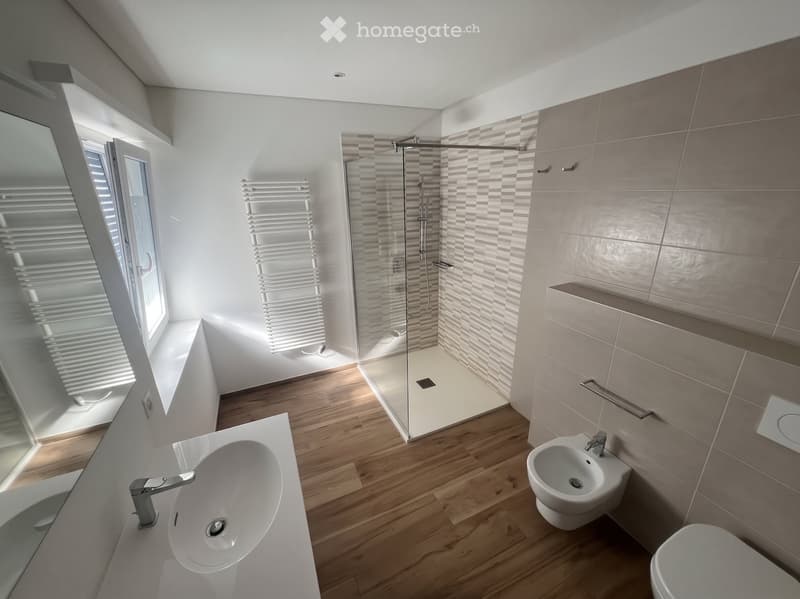 Modern bathroom with wooden flooring, white sink, toilet, bidet, and a glass-enclosed shower area. Features a radiator and a window with blinds.