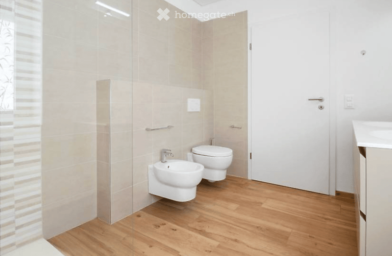 Modern bathroom with wooden flooring, white tiles, a toilet, a bidet, and a sink. Features a glass shower enclosure and a white door.