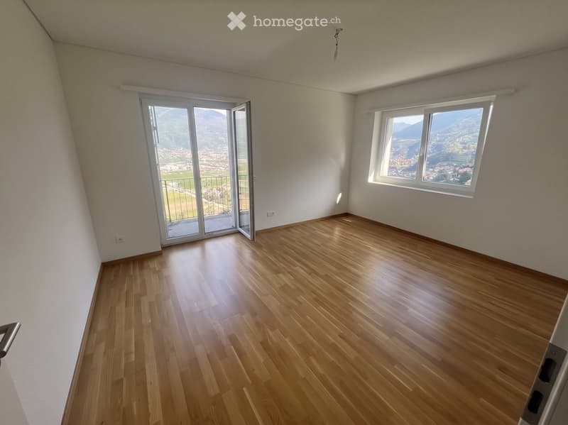 Empty room with wooden flooring, large windows offering a scenic mountain view, and a sliding glass door leading to a balcony.