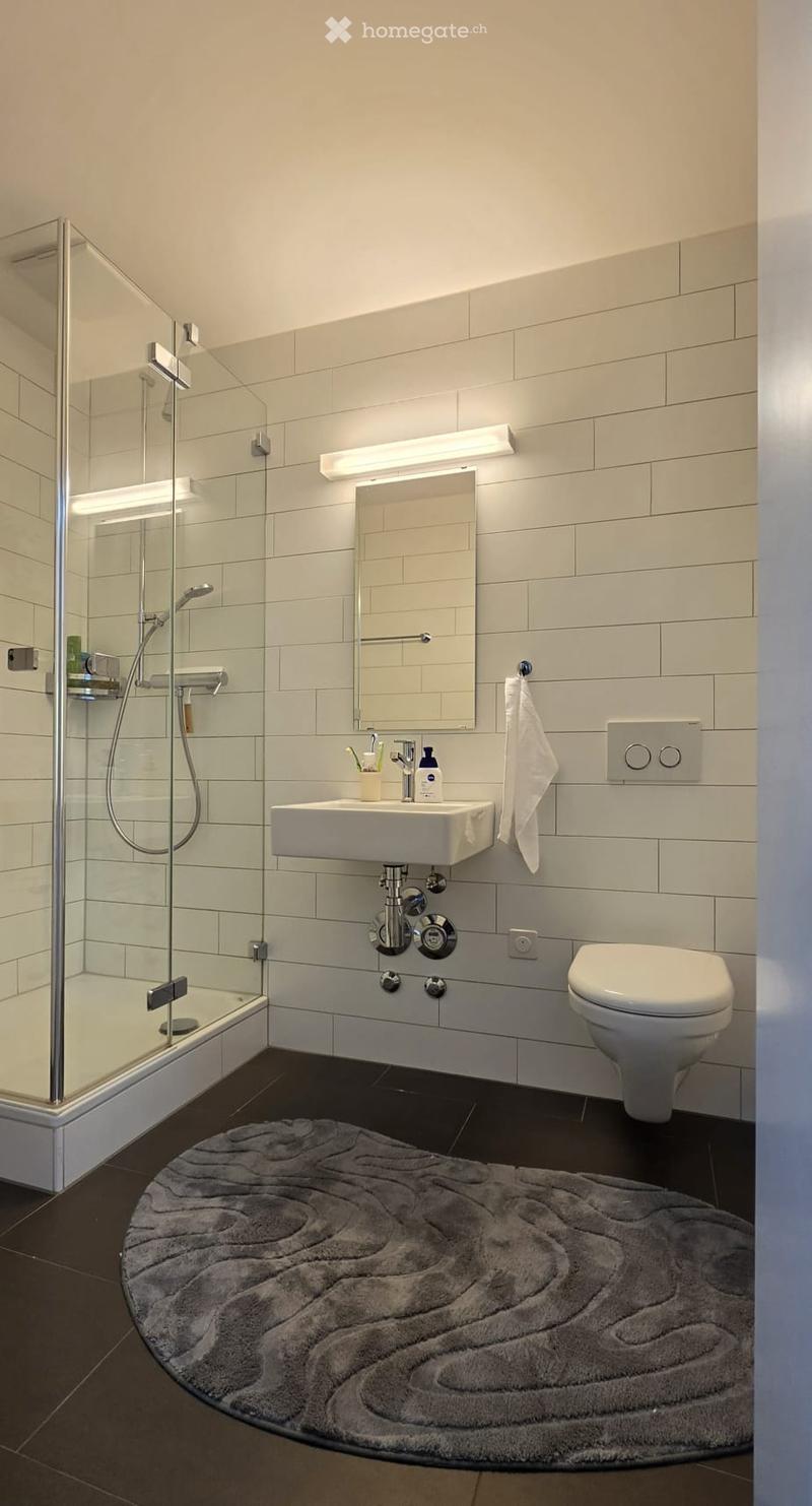 Modern bathroom with white tiles, a glass-enclosed shower, a white sink with a mirror, a toilet, and a soft grey round rug on the dark floor.