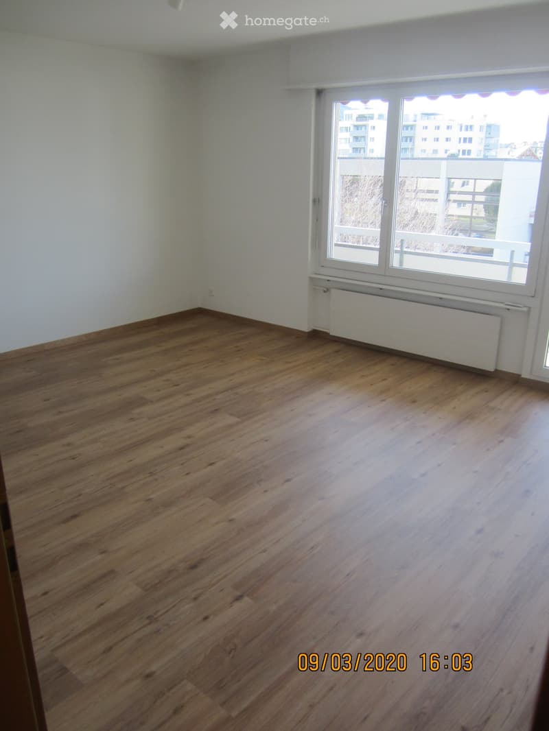 Empty room with wooden flooring, white walls, and a large window with a view of a building and balcony.
