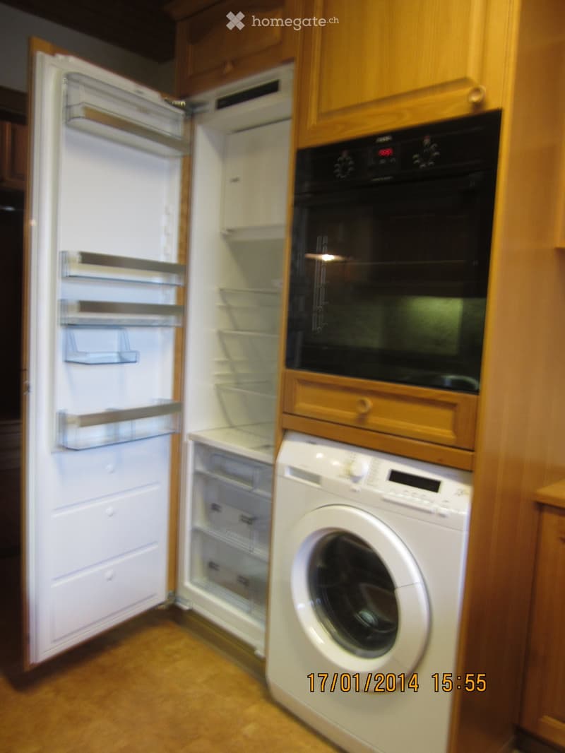 Compact kitchen area with wooden cabinets, open refrigerator, oven, and washing machine.