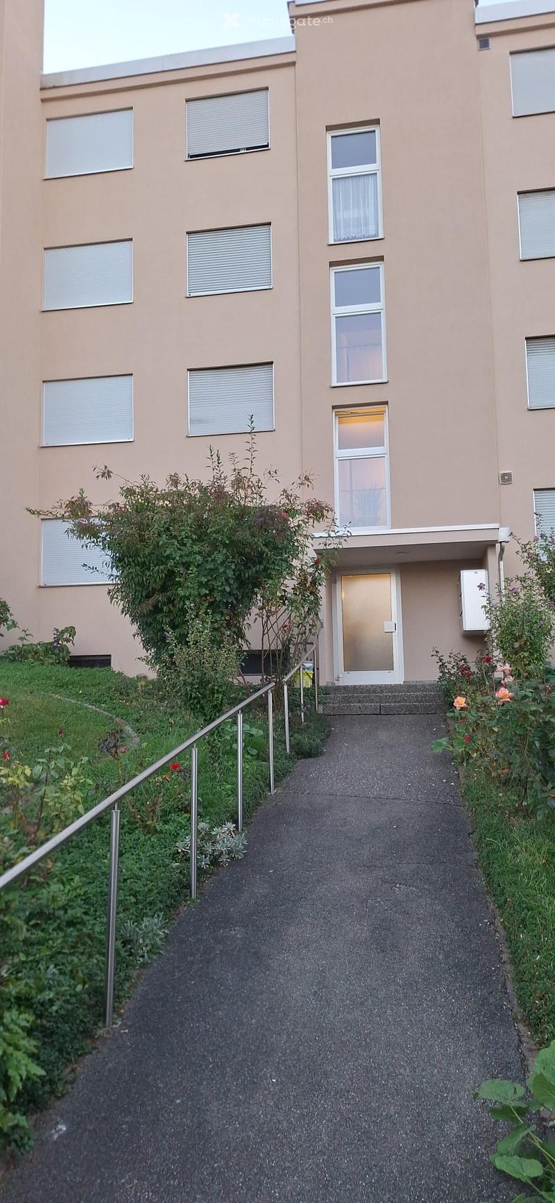 A two-story residential building with beige walls, multiple windows, and a small garden area with plants and a metal handrail leading to the entrance.