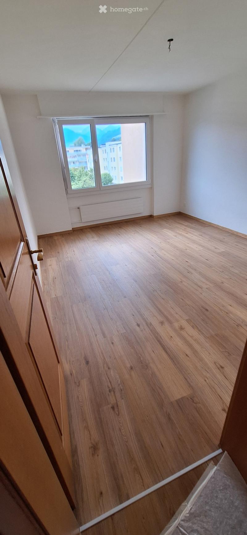 Empty room with wooden flooring, white walls, and a large window with a view of buildings and trees outside.