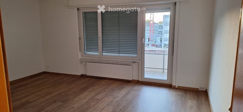 Empty room with wooden flooring, white walls, large window with blinds, and a sliding glass door leading to a balcony.