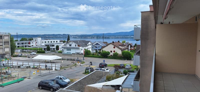 Balcony overlooking a street with parked cars, a white tent, and a distant view of a lake and mountains.
