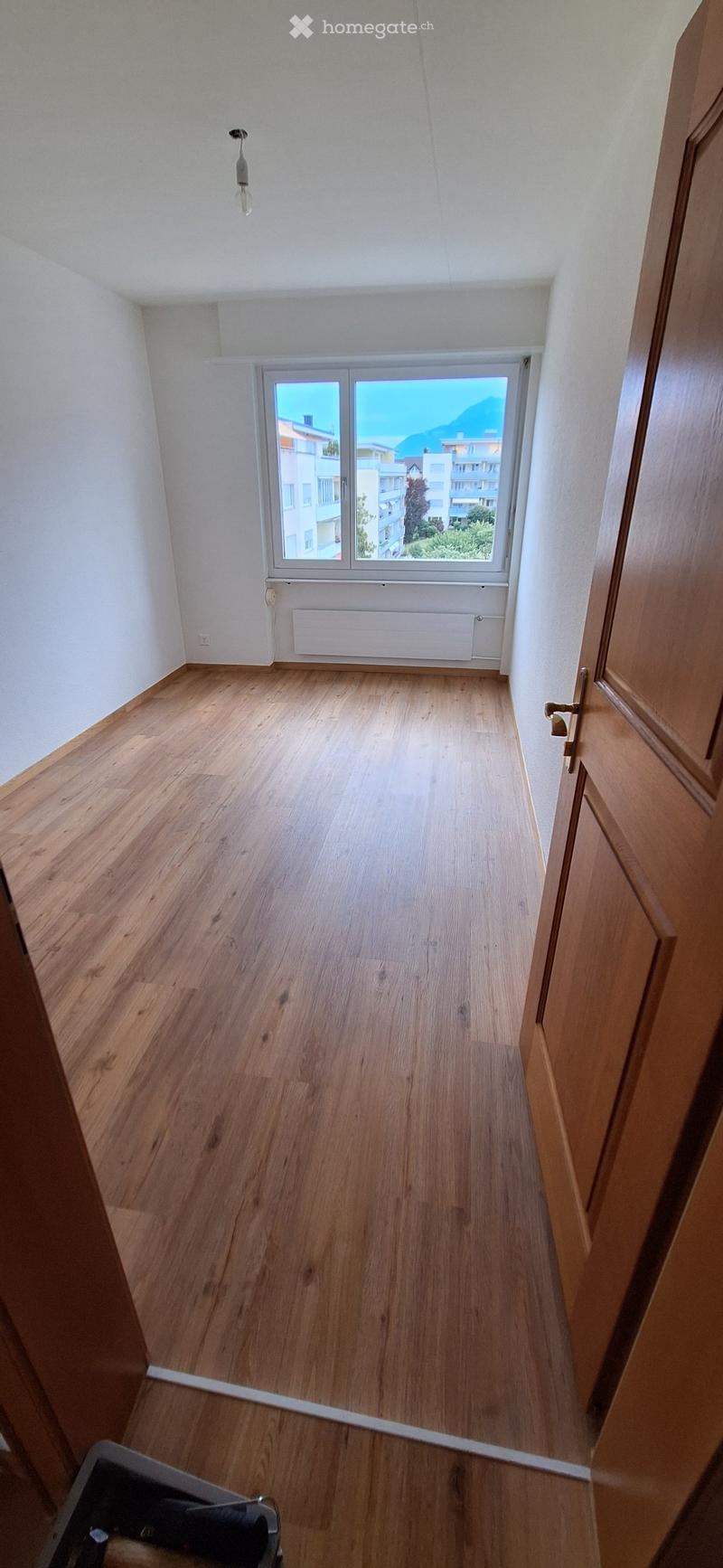 Empty room with wooden flooring, white walls, and a large window offering a view of buildings and mountains.
