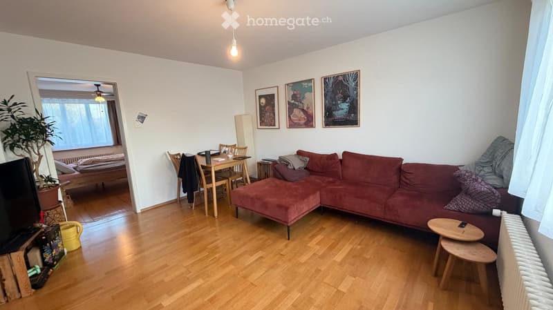 A cozy living room with a red sectional sofa, a small wooden dining table with chairs, a potted plant, and several picture frames on the wall. The room features wooden flooring and a hanging light fixture.