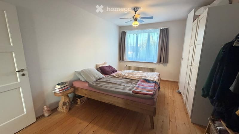 Cozy bedroom with wooden flooring, a single bed with pillows and blankets, a small wooden stool with books, a radiator, and a wardrobe with clothes hanging on it.