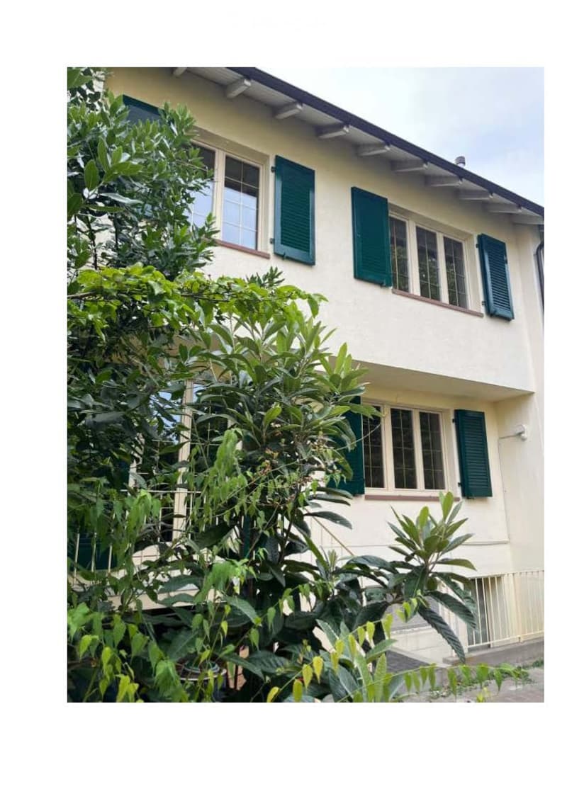 Two-story house with light yellow walls, green shutters, and multiple windows partially obscured by lush green foliage.