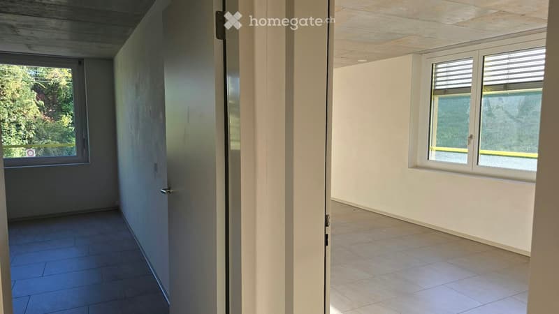 Empty room with tiled floor, white walls, and two windows with blinds. One door is open, revealing a view of greenery outside.