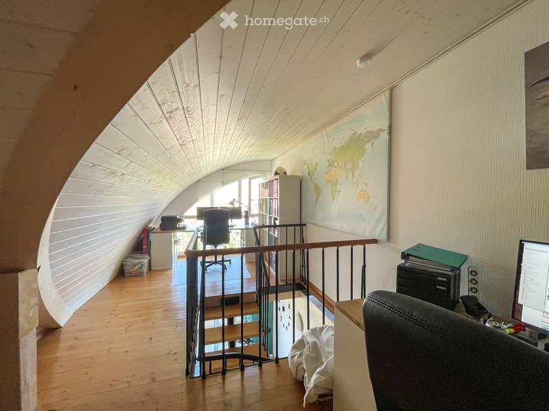Office space with wooden flooring, slanted ceiling, and a railing overlooking a lower level. Includes a desk, chair, laptop, bookshelf, and a world map on the wall.