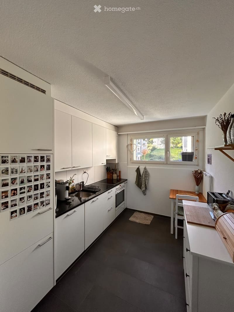 Modern kitchen with white cabinets, black countertops, and stainless steel appliances. Features a refrigerator with photo collage, a small dining area with a table and chairs, and a window overlooking a green area.