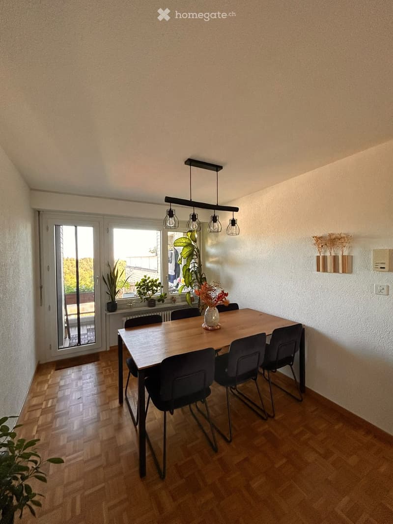 Dining room with wooden floor, large window, dining table with chairs, vase with flowers, and wall-mounted shelf with dried plants.