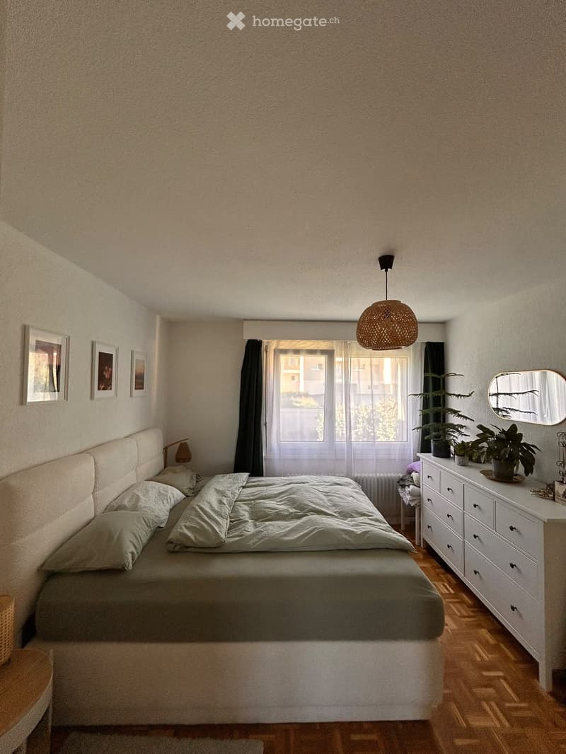 Cozy bedroom with a large bed, nightstand, lamp, mirror, and plants. The room features wooden flooring and a large window with curtains.